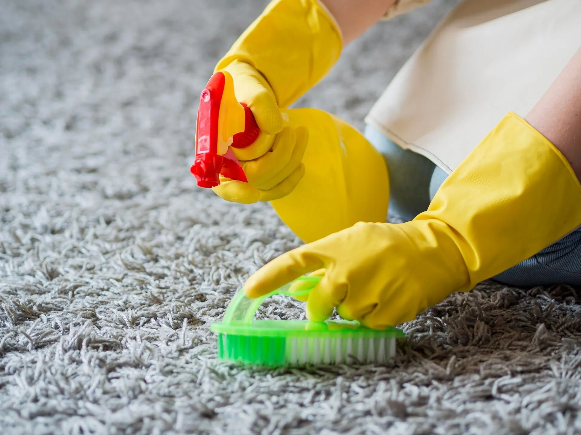 A person wearing yellow gloves is cleaning a carpet with a spray bottle and a brush.