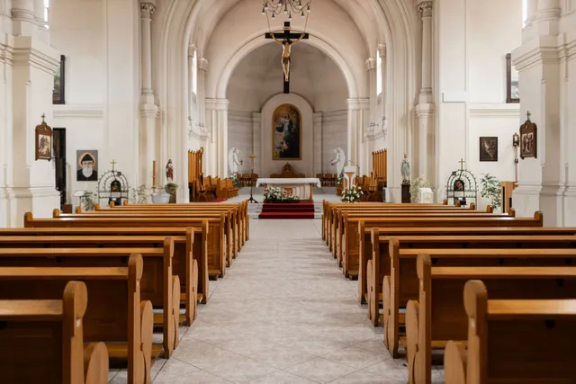 The inside of an empty church with wooden benches and a cross on the altar.